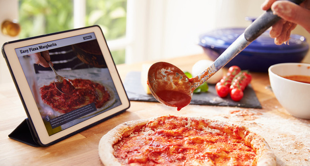 Tomatensoße wird mit einer Kelle auf einer Pizza verteilt. Dahinter steht ein Tablet, auf dem eine Rezepte-App läuft. Bild: Monkey Business / Fotolia.com